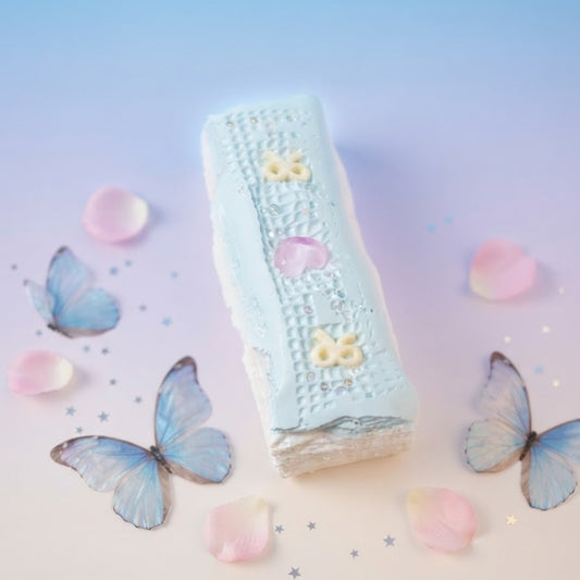 A rectangular pastel blue cake or pastry with white icing and small yellow decorative details, styled on a soft blue-to-pink gradient background with blue butterflies, pink petals, and small star confetti.