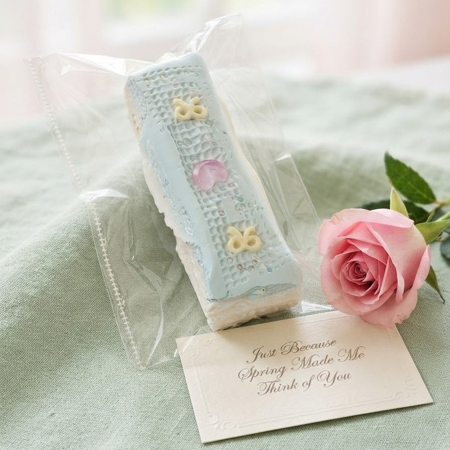 A long rectangular pastel cake wrapped in clear cellophane packaging, placed on a pale green fabric surface beside a pink rose and a small handwritten note.