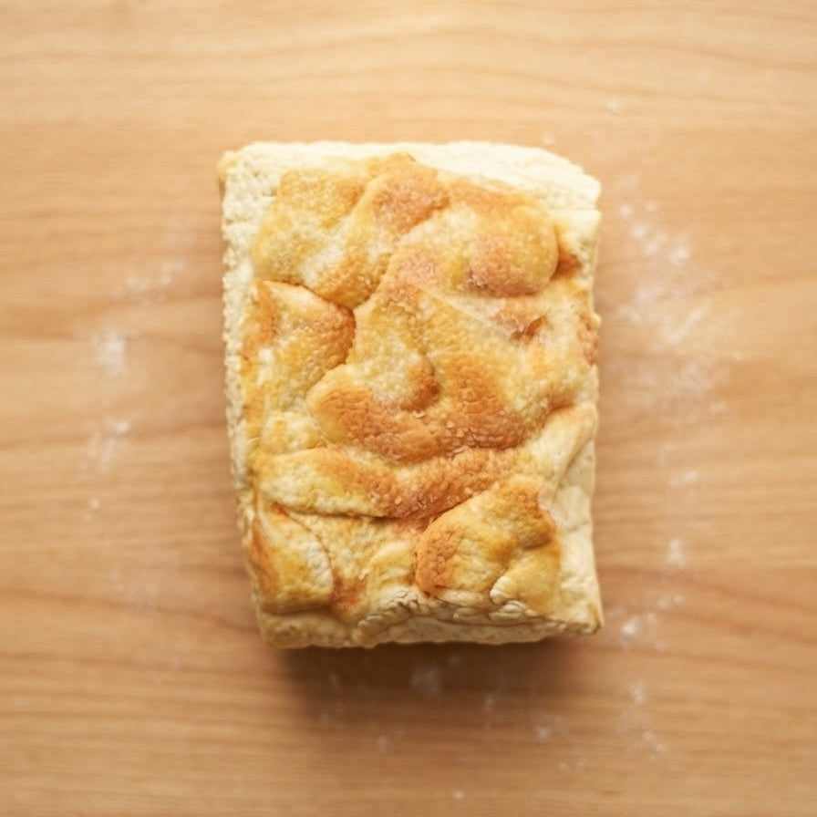 A rectangular piece of golden-brown baked bread or pastry with a textured, swirled top crust, placed on a light wooden surface with a few scattered flour particles.