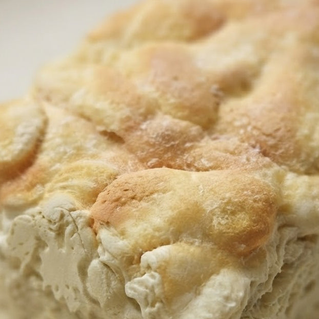 A close-up shot of a freshly baked, golden-brown focaccia bread with a dusting of flour on a light background.