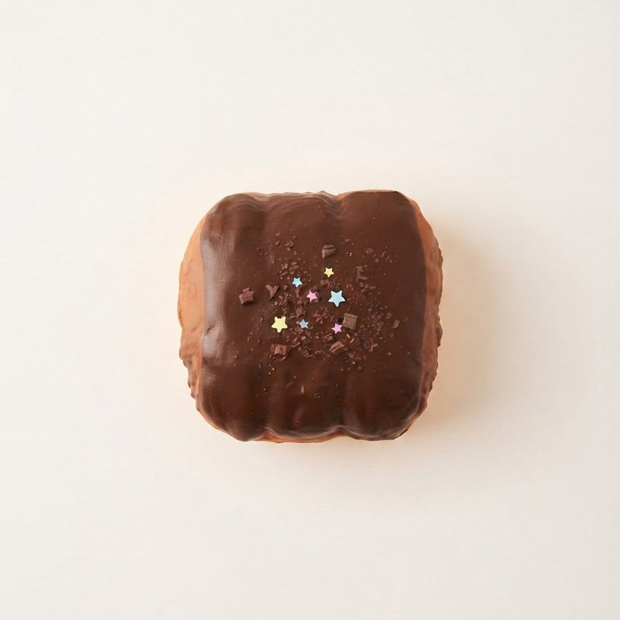 A chocolate-covered pastry or donut with a glossy dark brown glaze, topped with small multicolored star sprinkles and chocolate crumbs, centered on a plain off-white background.