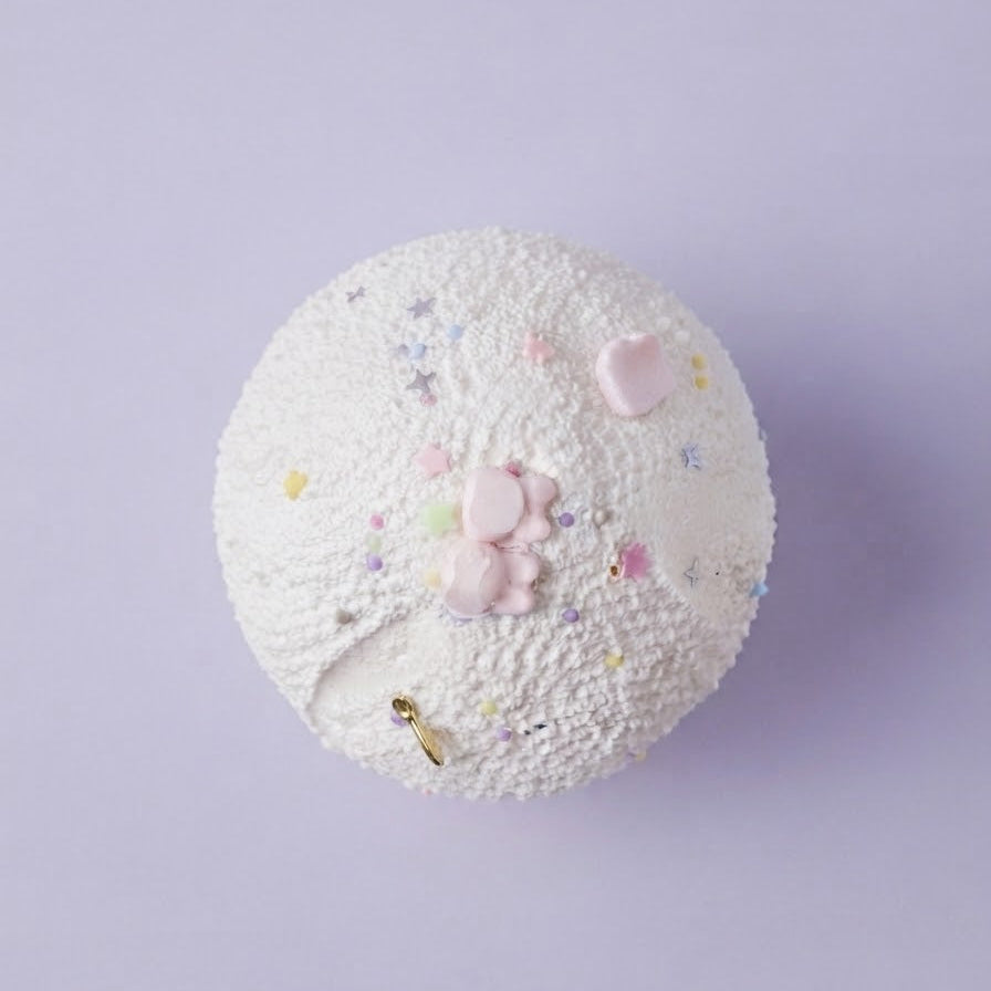 A round, textured white cupcake topper or decorative cake decoration with pink frosting details, small pastel sprinkles, and a tiny gold key accent, shown on a solid light purple background.