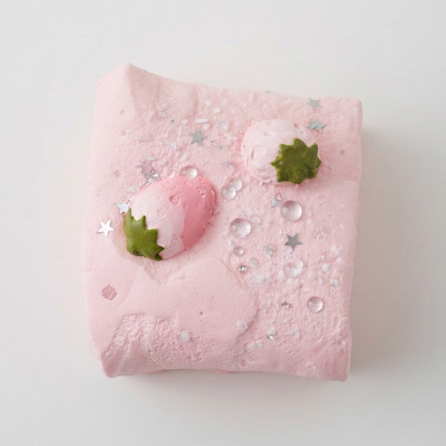 A pink square bath bomb or soap bar decorated with two small green strawberry tops, silver star sprinkles, and clear rhinestones, photographed on a clean white background.
