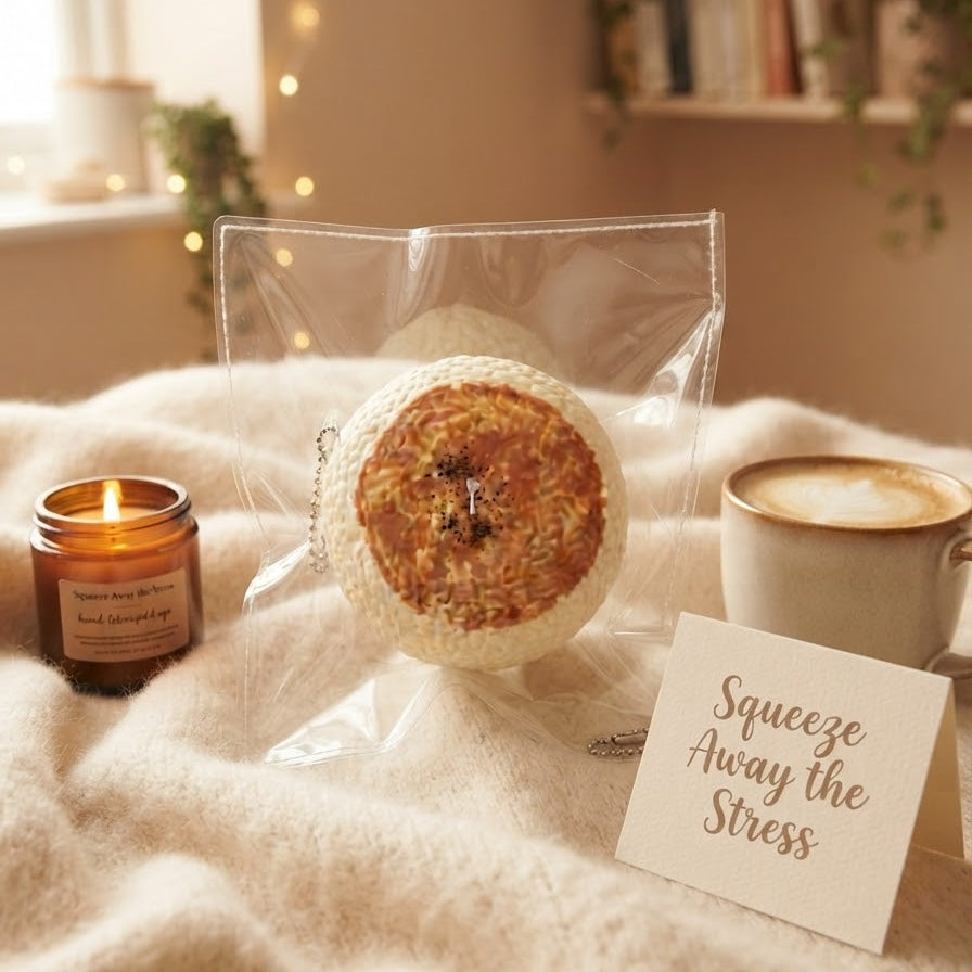 A round bath bomb in a clear plastic bag sits on a cream knit blanket, styled with a lit candle, a cup of coffee, and a card that reads 'Squeeze Away the Stress'.