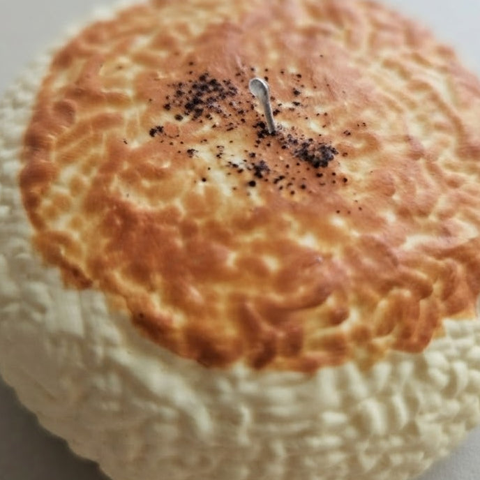 A round, textured novelty candle shaped like a baked bread or pastry, with a golden-brown top and a small metal wick, photographed on a light tabletop.