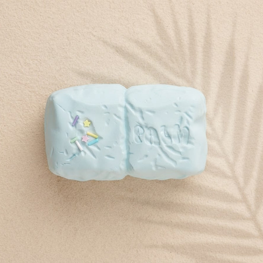 A light blue double-bar soap block with embossed lettering and a small colorful unicorn illustration, displayed on a beige textured surface with palm leaf shadows.