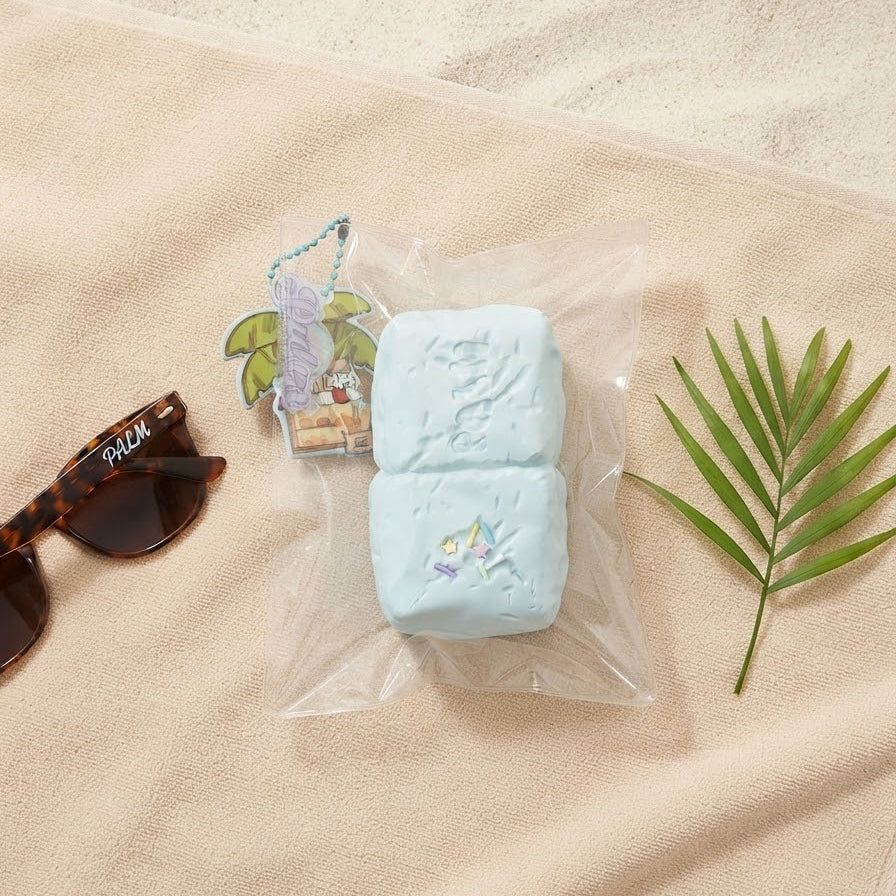 A light blue rectangular bath sponge with a small tropical illustration, packaged in a clear plastic bag and placed on a beige towel with sunglasses and a green palm leaf nearby.