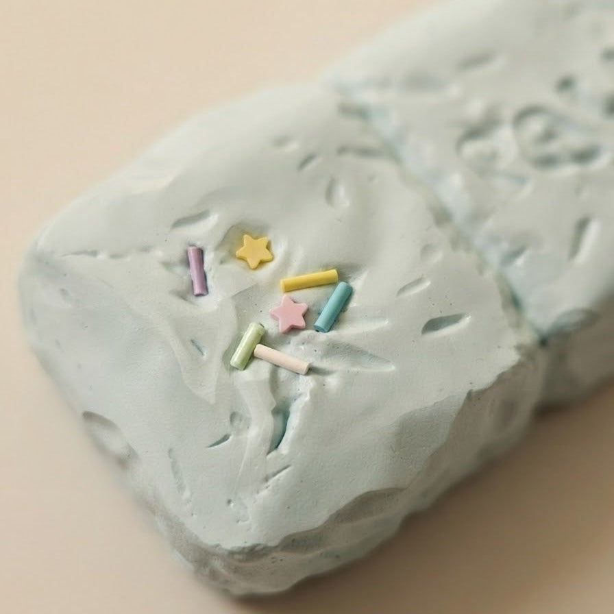 A close-up of a pale mint green textured soap bar decorated with small pastel sprinkles, set against a solid beige background.