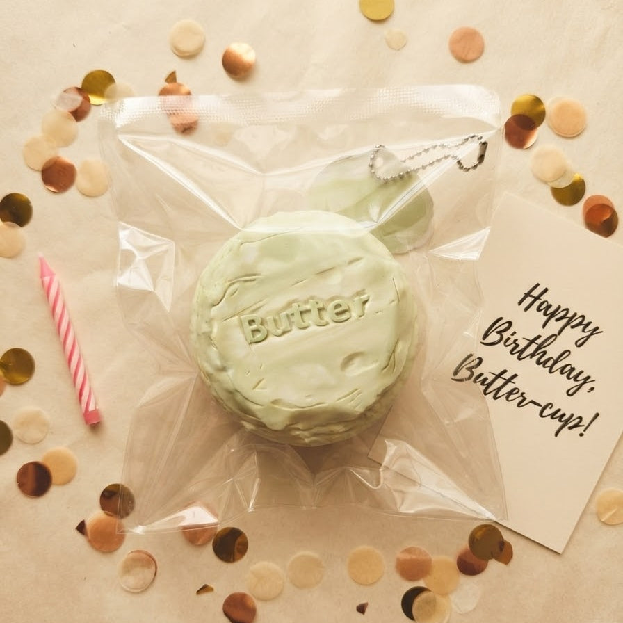 A light green, cheese-like squishy toy with the word 'Butter' embossed on the front, packaged in a clear plastic bag with a keychain loop. The package is arranged on a beige paper background with scattered gold and bronze confetti, a pink striped candle, and a handwritten card that reads 'Happy Birthday, Butter-cup!'.