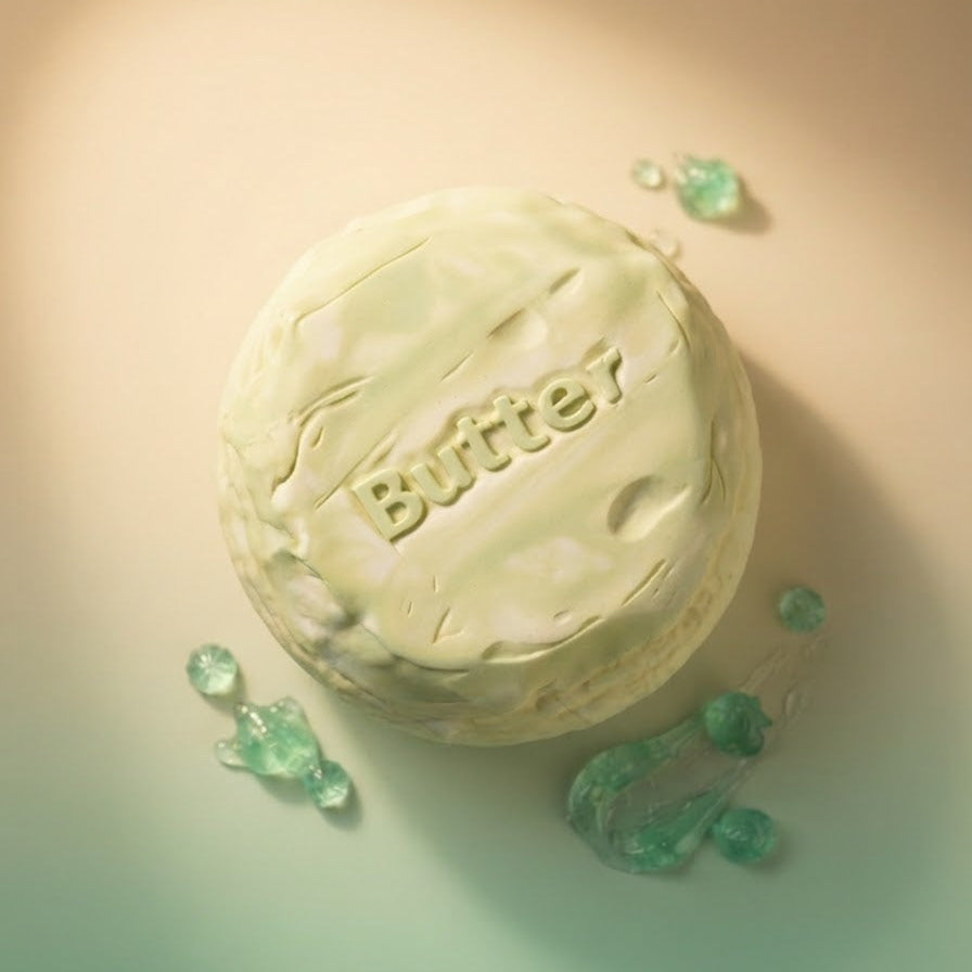 A round, off-white bar of soap with the brand name 'Butter7' embossed on top, styled on a warm beige background with green translucent decorative droplets.