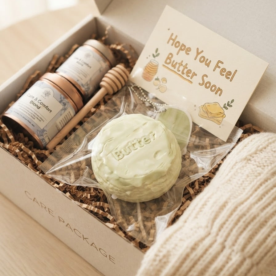 A gift box containing a pale green butter puck in clear wrapping, two small jars, a wooden honey dipper, and a card that reads 'Hope You Feel Butter Soon.' The box is placed on a light wood surface with a cream knit fabric beside it.