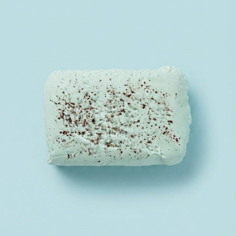 A rectangular bath bomb with a textured, frosted off-white surface and dark brown cocoa powder sprinkles, shown on a solid light blue background.