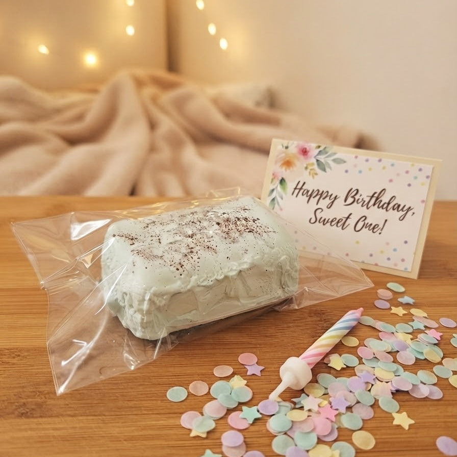 A packaged frosted cake-shaped item on a wooden table with a small card reading 'Happy Birthday Sweet One!' and pastel confetti scattered around. A real indoor setting with warm string lights and a blanket is visible in the background, so this is Lifestyle rather than Product/Packaging.