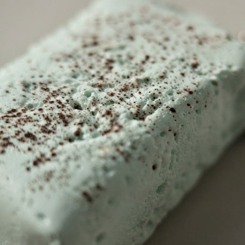 A close-up of a rectangular, light mint-green bath bomb or soap bar with a textured, porous surface and dark brown speckles, shown on a neutral gray background with a chain partially visible on the right.