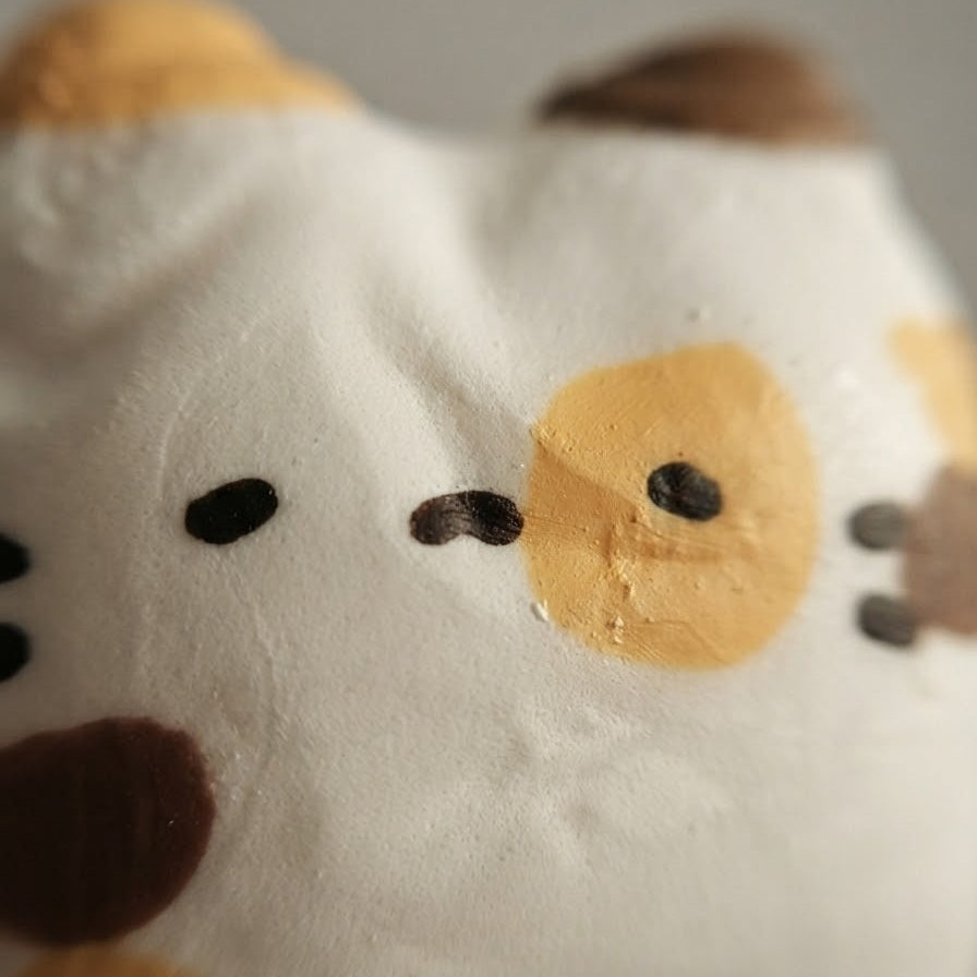 A close-up of a white plush toy or cushion with a cat face design, featuring yellow and brown patches and small black eyes and nose details.