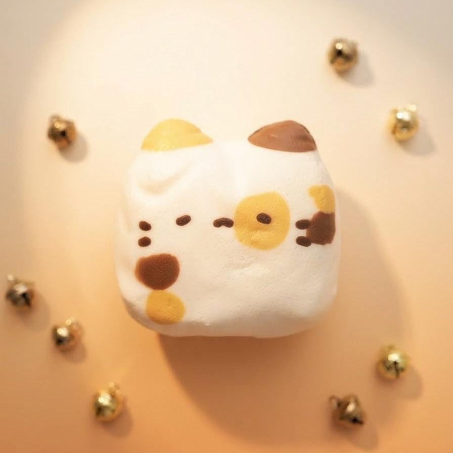 A white plush cat-shaped pillow with yellow and brown embroidered facial features, photographed from above on a warm peach background with scattered small gold bells.