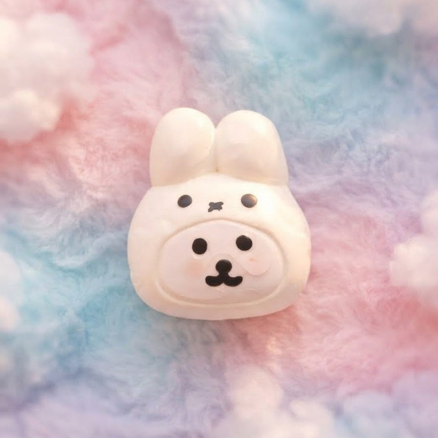 A small white bunny-shaped accessory with black facial features, photographed against a soft pastel cotton candy background of pink, blue, and white.