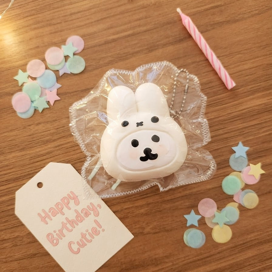 A cute white bunny-shaped keychain or charm with a black face, wrapped in clear cellophane, displayed on a wooden table with a pink candle, pastel confetti, and a 'Happy Birthday Cute!' tag.