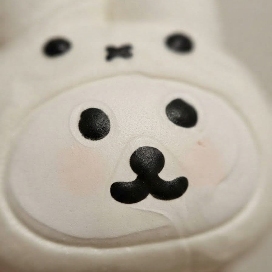 A close-up of a white plush toy or stuffed animal face with black embroidered eyes, nose, and mouth details, held against a soft blurred background.