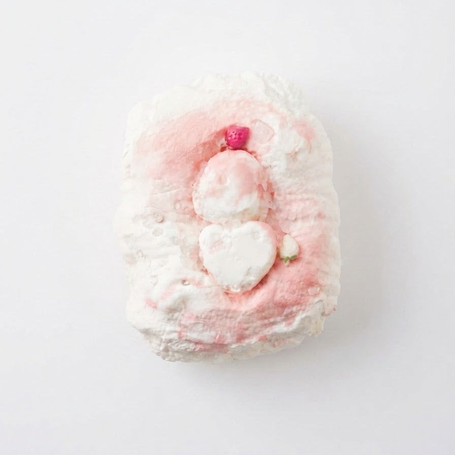 A single bath bomb with a textured, bubbly surface in white and soft pink, featuring a small pink heart detail on top, photographed from directly above on a clean white background.