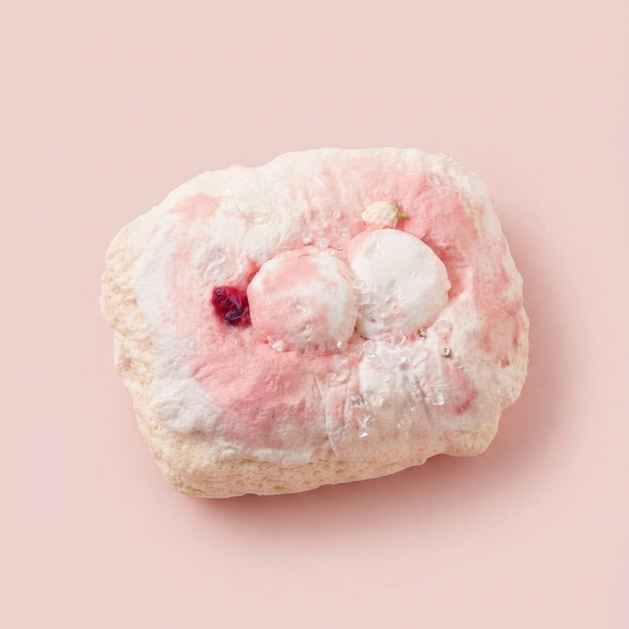 A pink and white marshmallow-themed pastry or baked good with a small dark red berry accent, photographed on a solid light pink background.