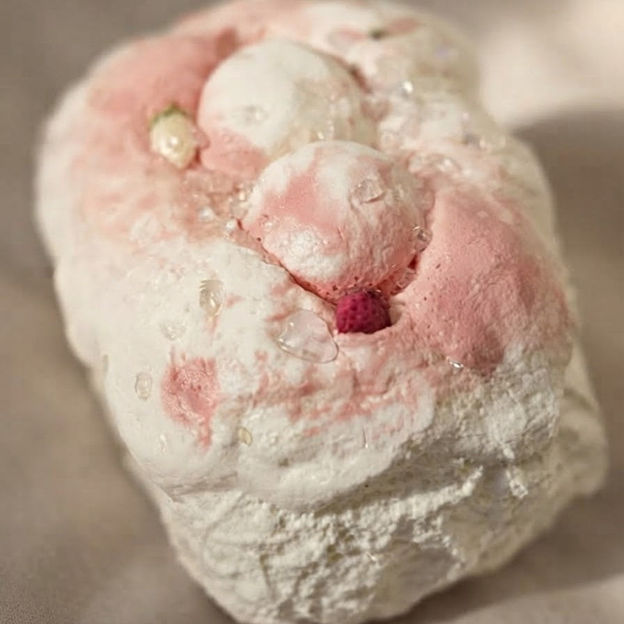 A close-up of a textured, organic-shaped product with a white and light pink surface, featuring raised bubble-like details and a small dark pink accent, resting on a neutral beige background.
