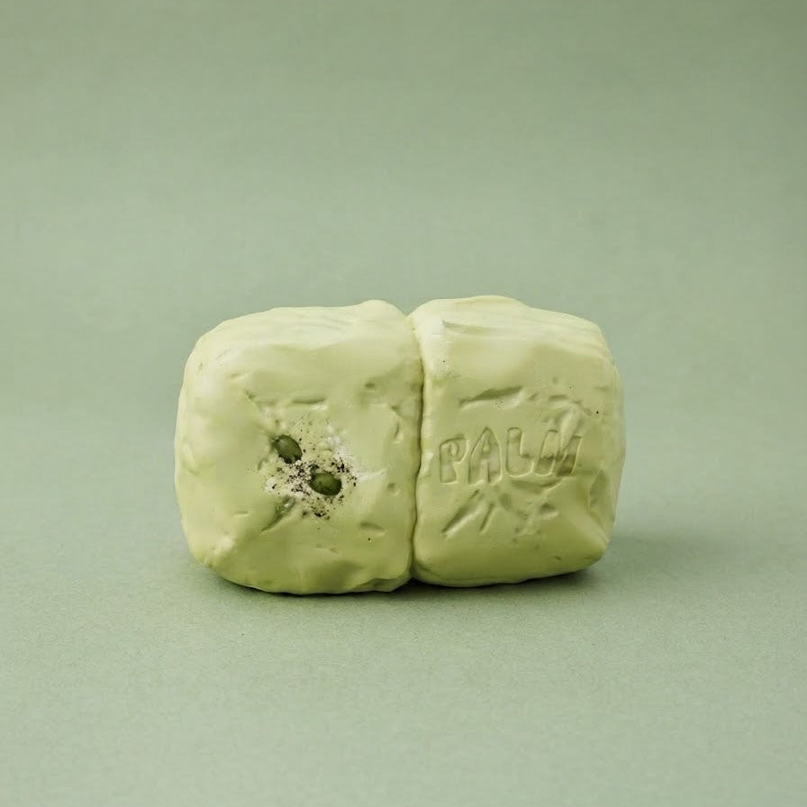 A small pale green clay or wax object with a rough, molded texture, standing upright on a smooth matching green background. The right side shows the embossed word 'PALM'.