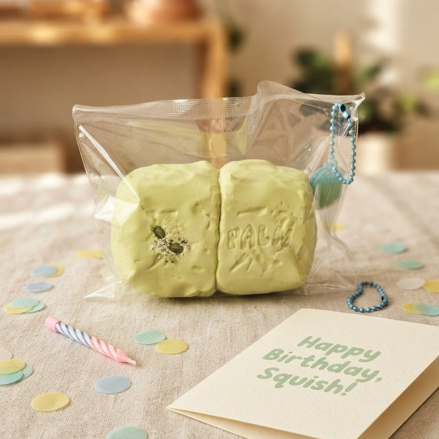 A pale yellow-green squishy toy shaped like two stacked cake slices, sealed in a clear plastic bag with a blue bead chain. It sits on a beige fabric surface with pastel confetti, a birthday candle, and a card that reads 'Happy Birthday, Squish!'.