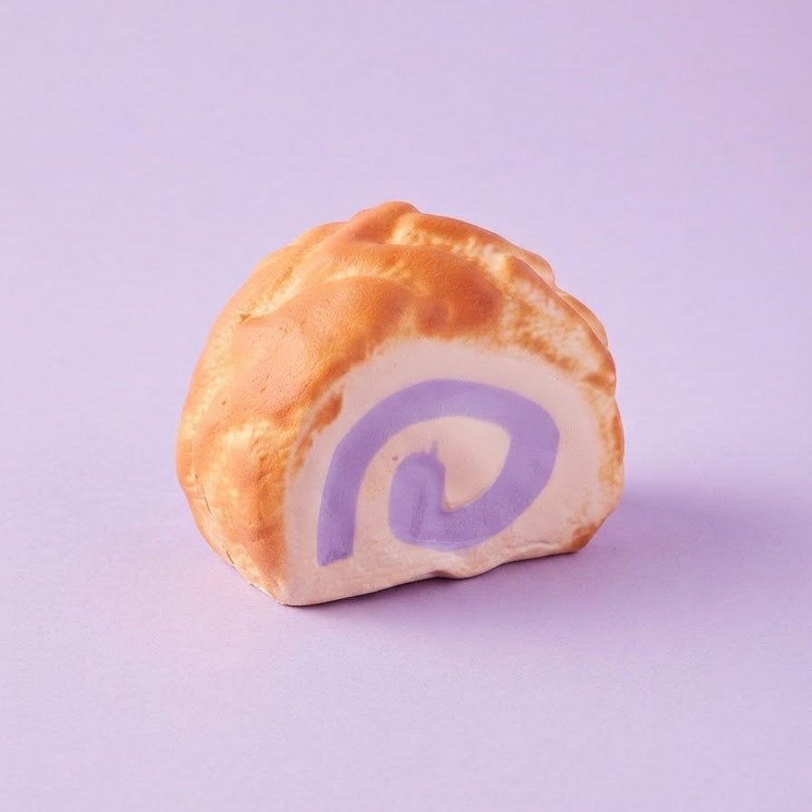 A novelty prank toy shaped like a pastry roll, featuring a golden-brown textured exterior and a purple spiral swirl inside, set against a solid lavender background.