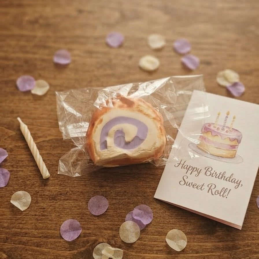 A wrapped sweet roll with a purple swirl sits on a wooden table beside a printed card that says 'Happy Birthday, Sweet Roll!' and a small birthday candle. Purple and white confetti is scattered around.