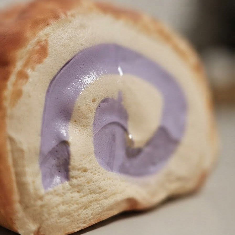 A close-up of a baked pastry roll with a golden-brown crust and a light beige filling swirled with a thick lavender-purple layer.