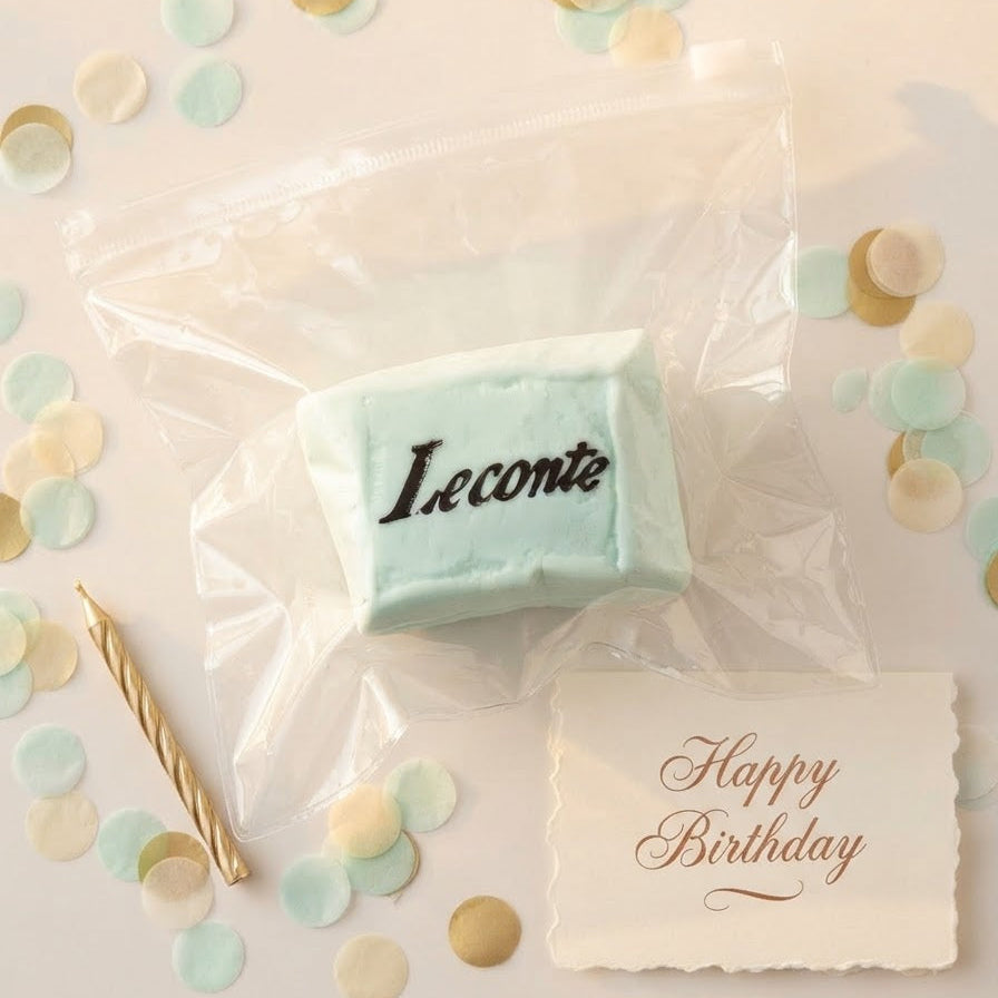 A light blue rectangular cake topper or favor piece with the name 'Leconte' in black script, sealed in a clear plastic bag and arranged on a cream surface with pastel confetti, a gold candle, and a 'Happy Birthday' card.