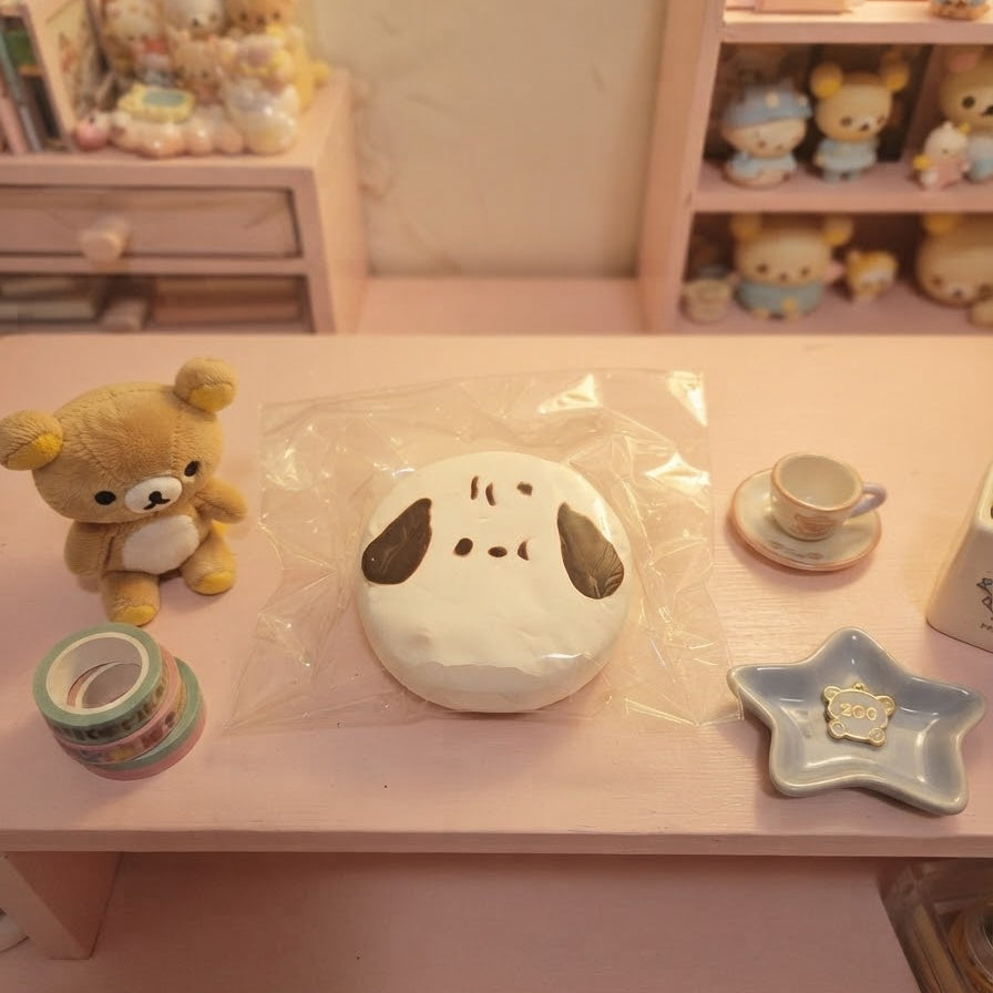 A small white dog-shaped squishy toy with dark brown ears and eyes, sealed in clear plastic, displayed on a pink table alongside a teddy bear, washi tape, a teacup, and a star-shaped dish in a miniature room setting.
