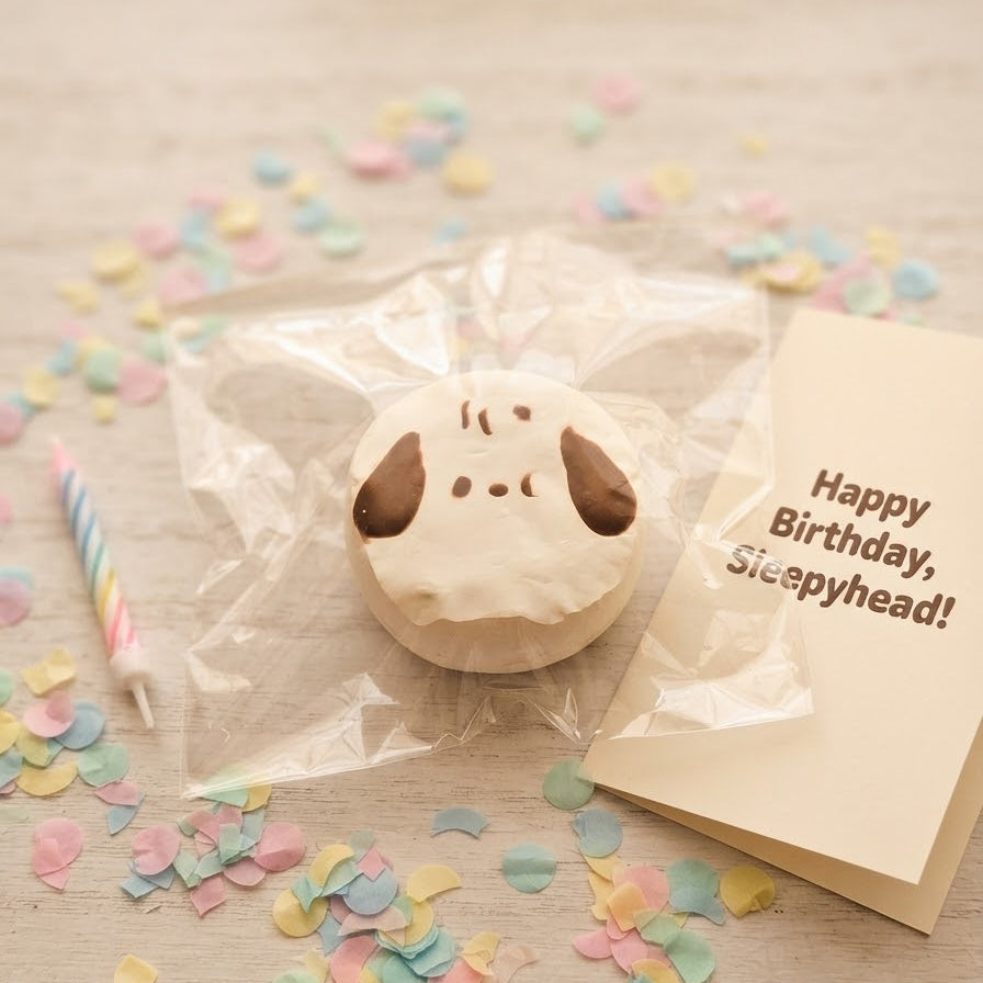 A cute dog-shaped squishy toy with white frosting and brown ears, wrapped in clear cellophane packaging next to a 'Happy Birthday, Sleepyhead!' greeting card and a striped candle, on a light wooden table scattered with pastel confetti.