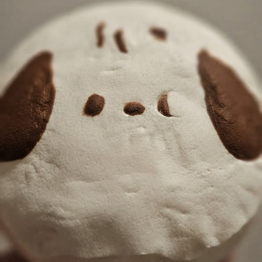 A close-up of a white plush toy or cushion with brown embroidered facial features, including two large floppy ears and small stitched eyes and mouth details.