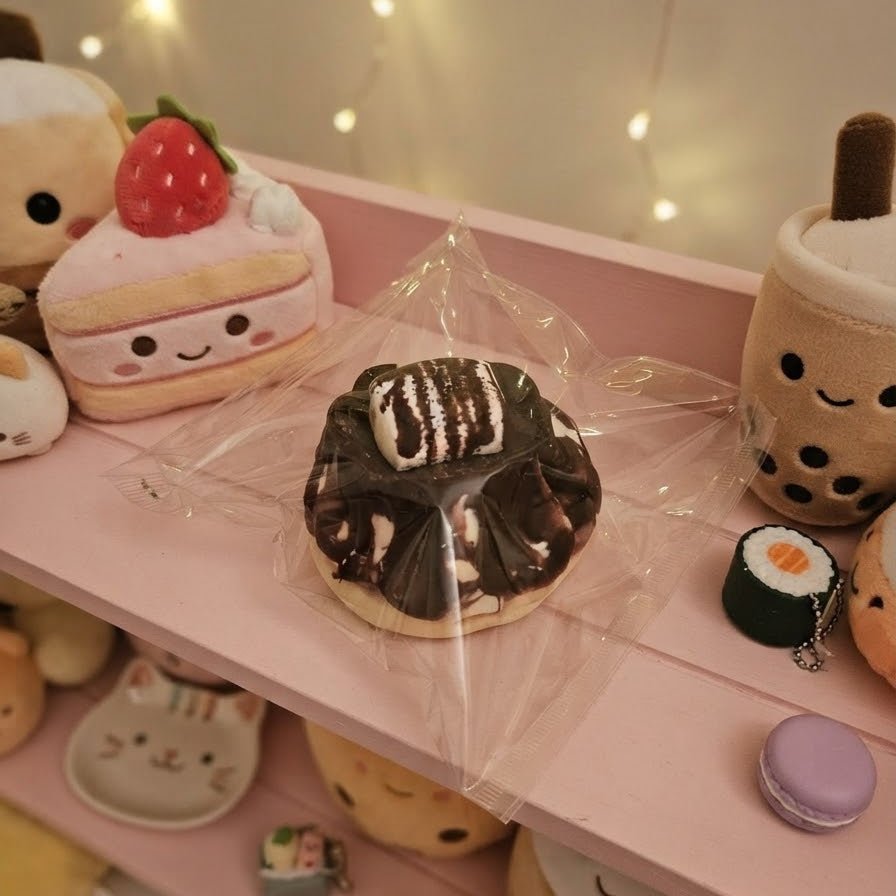 A chocolate donut-shaped squishy toy wrapped in clear plastic, displayed on a pink shelf among other cute plush food toys like a strawberry cake slice, boba cup, sushi roll, and macaron.