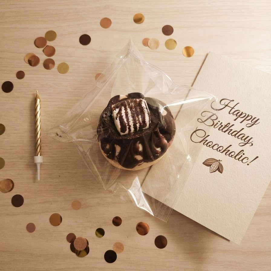 A chocolate-covered cupcake wrapped in clear cellophane sits on a light wood surface with scattered gold and bronze confetti, a gold candle, and a cream card that reads 'Happy Birthday, Checcohatic!'.