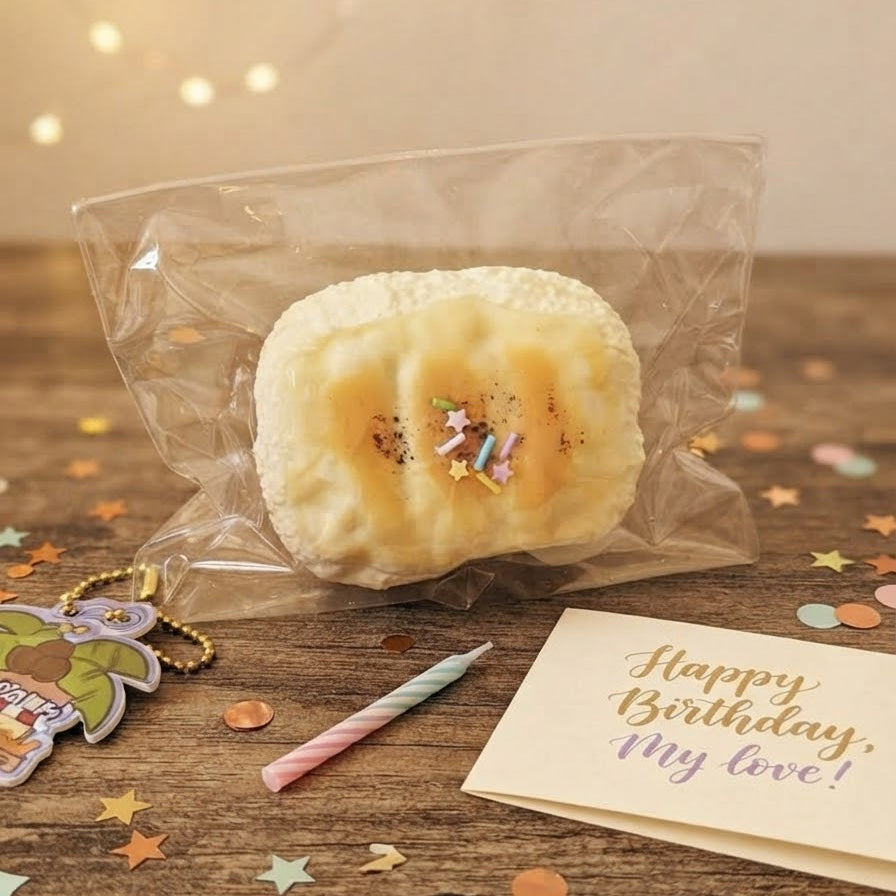 A packaged baked good, likely a novelty cake or pastry, sealed in a clear plastic bag and placed on a wooden tabletop with confetti, a candle, and a 'Happy Birthday, my love!' card nearby.