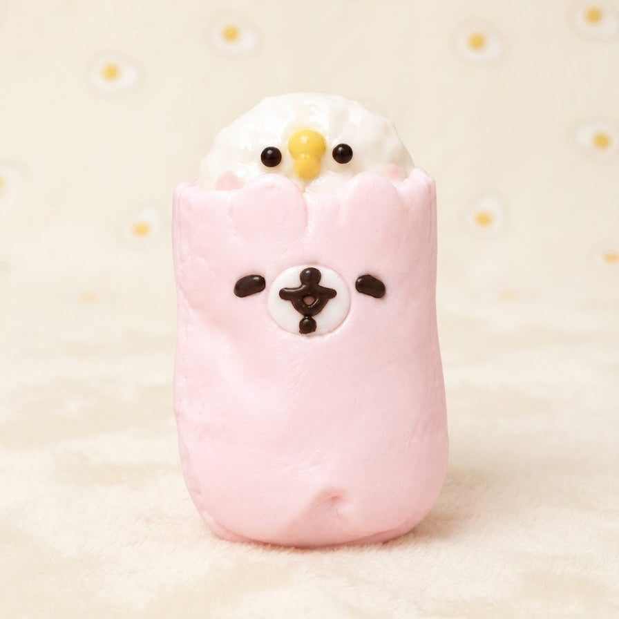 A cute pink and white character figurine resembling a fluffy bird or chick wrapped in a pink shell, with small black eyes, a yellow beak, and a white face patch, set against a warm cream background with small daisy patterns.