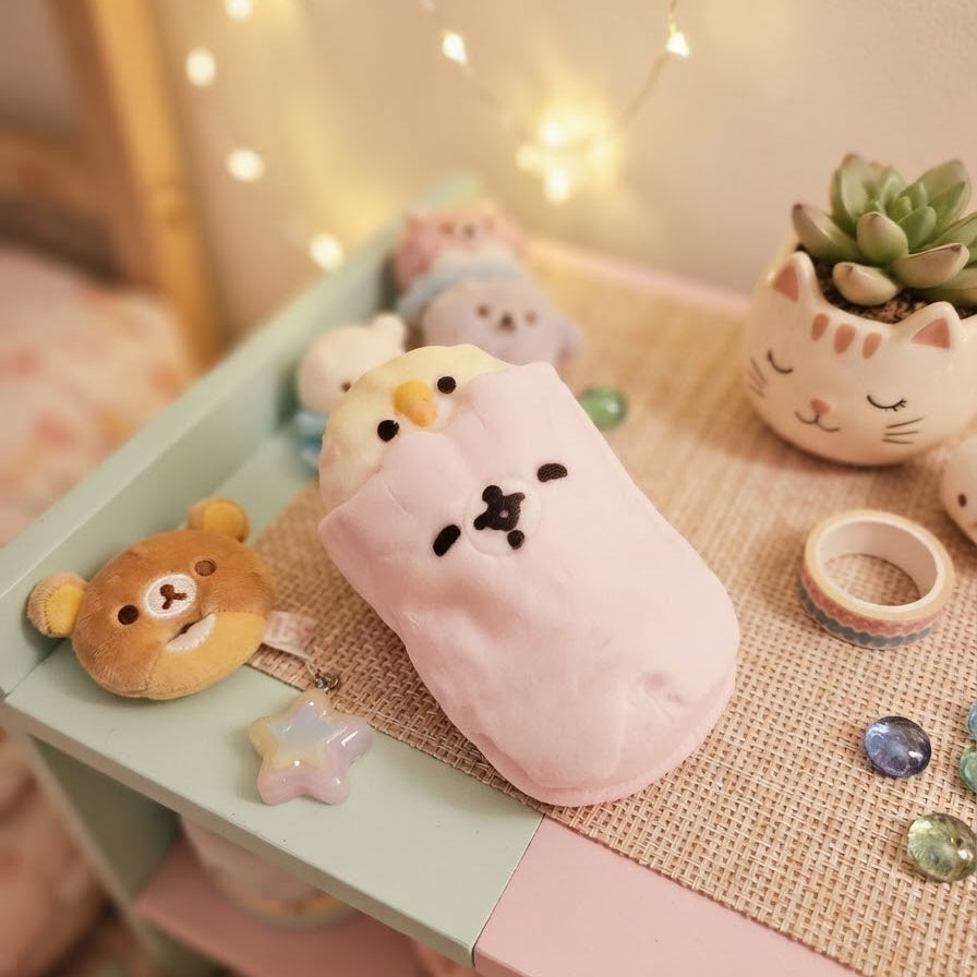 A pink plush pouch with a cute cartoon face design, displayed on a pastel-colored shelf alongside other kawaii accessories, including a small bear plush, a succulent in a cat-shaped pot, and decorative gems.