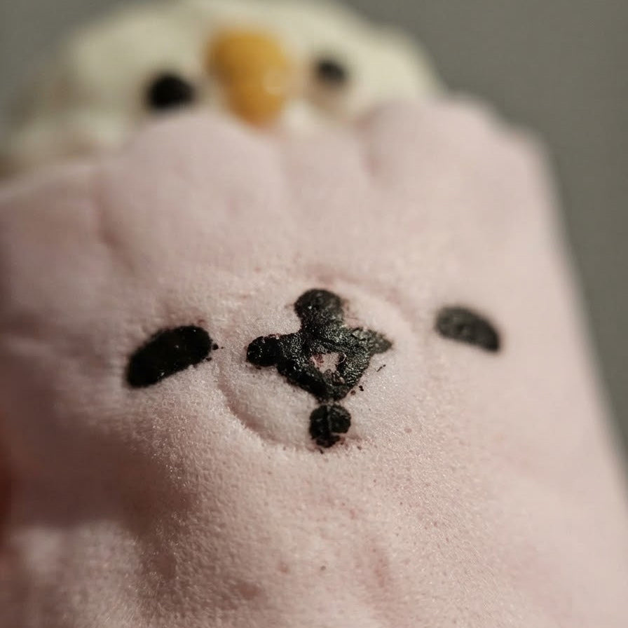 A close-up of a plush toy or stuffed animal in pale pink, showing embroidered black facial features and part of a white head with a small yellow accent against a soft gray background.