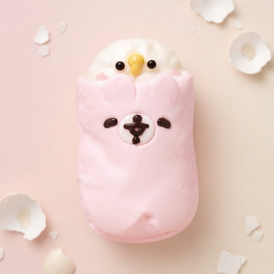 A pink and white fluffy cake or dessert shaped like a cute bird wrapped in a pink blanket, with a small yellow beak and dark facial details, photographed on a soft pink background with scattered white shell pieces.