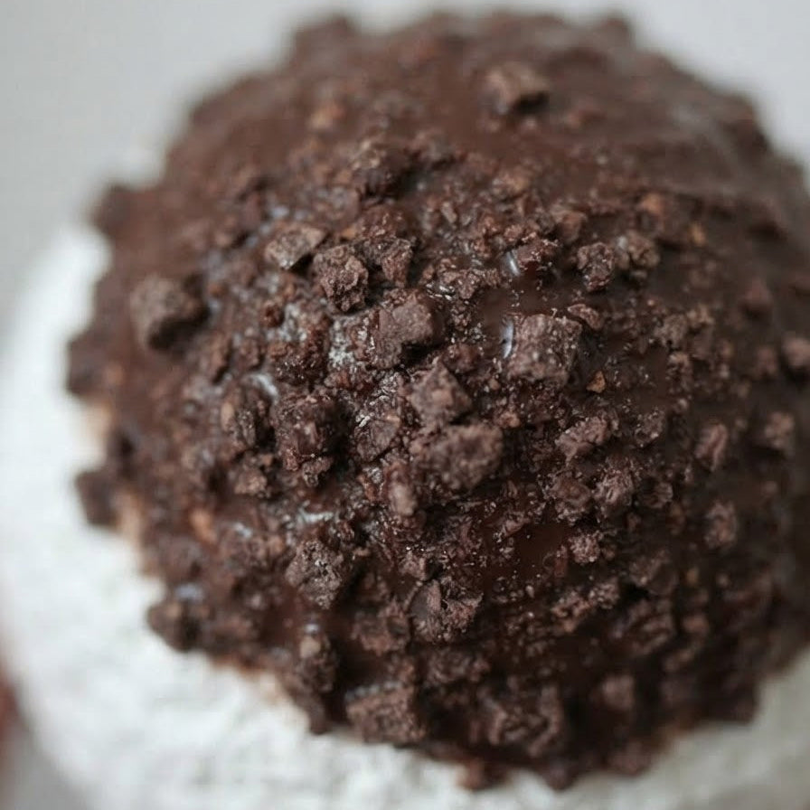 A close-up of a dessert, likely a scoop of vanilla ice cream topped with a glossy chocolate glaze and crushed cookie crumbs, set against a soft light gray background.