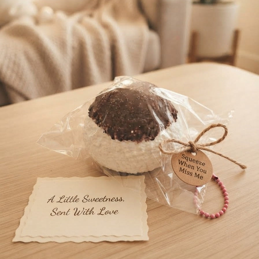 A small round novelty item wrapped in clear cellophane sits on a wooden table, with a handwritten note reading 'A Little Sweetness, Sent With Love' and a pink beaded tag attached. The background shows a softly blurred living room scene with a sofa and plant.