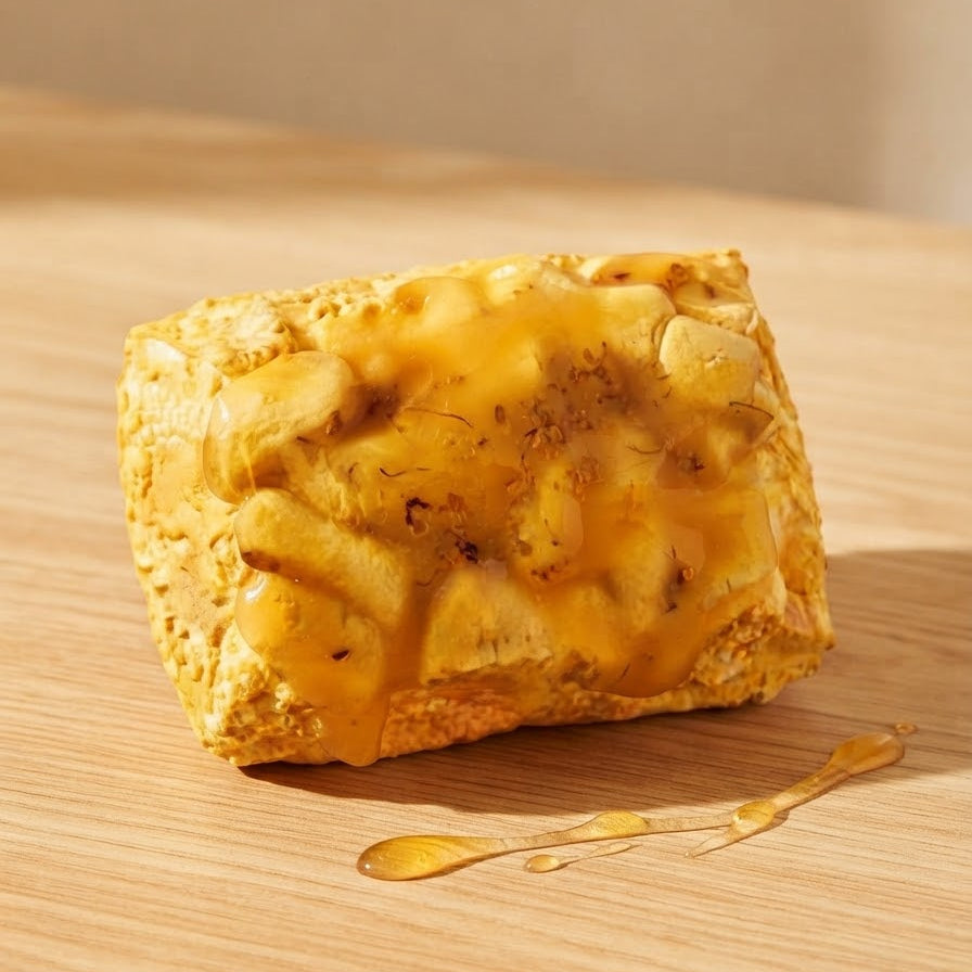 A square golden-brown baked good, possibly a cheese bread or savory pastry, topped with melted yellow cheese and drizzled with honey, resting on a light wooden surface.