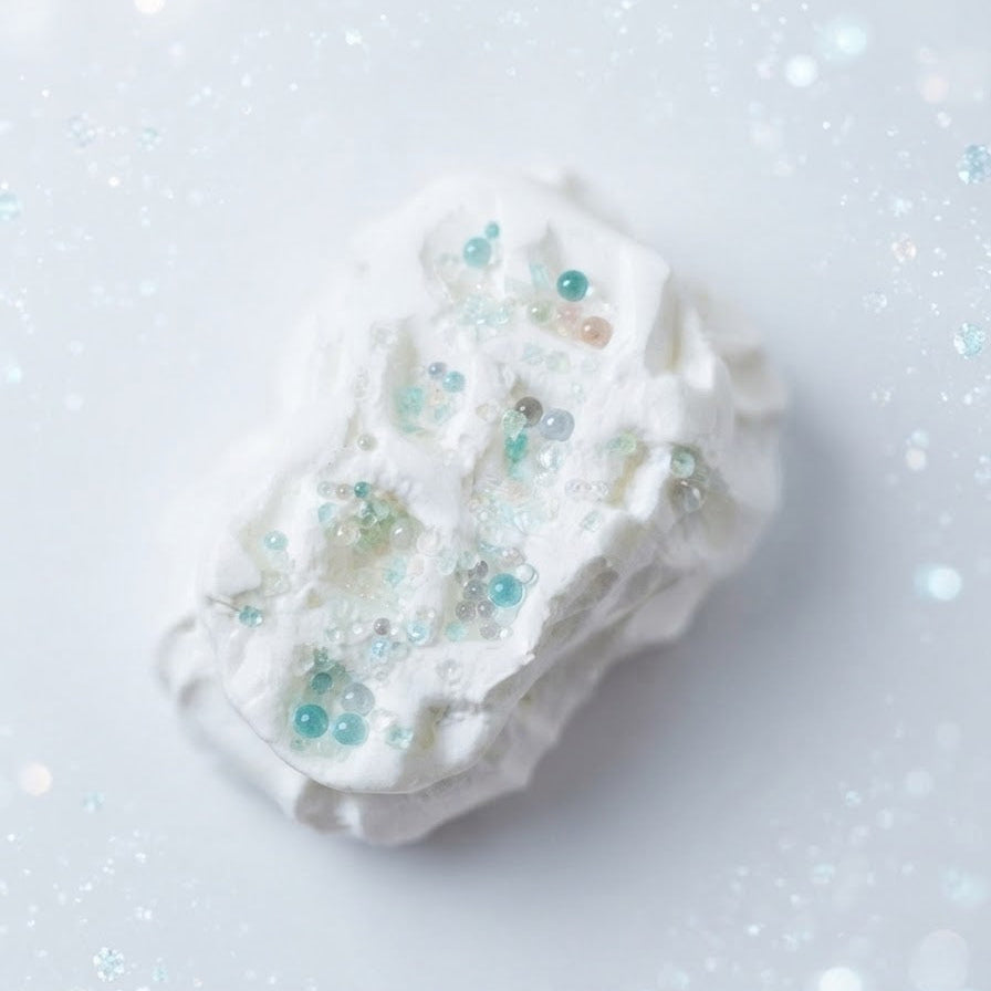 A white, textured foam or sponge-like product with scattered light blue and clear glitter beads, photographed on a soft white background with subtle bokeh sparkles.
