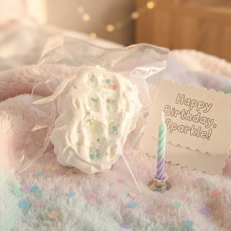 A white textured squishy toy with pastel glitter accents, wrapped in a clear plastic bag and placed on a fluffy pastel blanket next to a 'Happy Birthday, Sparkle!' card with a small candle.