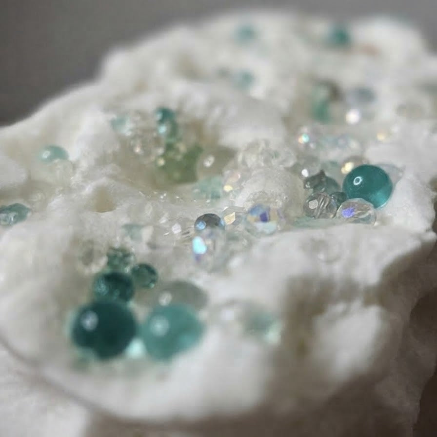 White textured fabric embellished with scattered teal and aqua beads, clear rhinestones, and iridescent sequins, shown in a close-up shot.