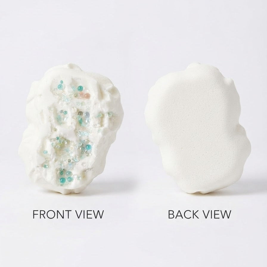 A white, textured, organic-shaped cosmetic sponge shown from both the front and back on a clean white background. The front view reveals small embedded glitter pieces in light blue, green, and gold.