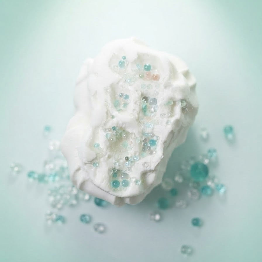 A white, textured bath bomb or soap piece with embedded clear and light blue beads, resting on a soft mint green background with scattered decorative beads around it.